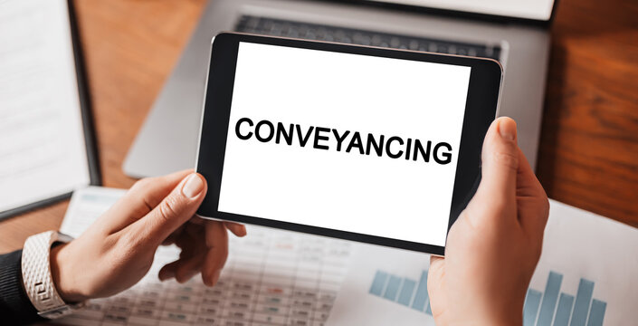 Man hands holding tablet with text Conveyancing at workplace. Businessman working at desk with documents