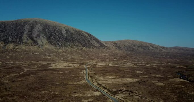 4k aerial footage of single track road through Glen Etive. No traffic on this road, A82 in distance with traffic. Mountains and Rannoch Moor background. Cloudless blue sky.