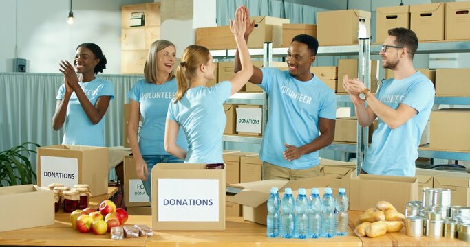 Joyful happy diverse Caucasian and African American men and women volunteers standing in social organization warehouse clapping hands and giving high five. Donations, charity work, volunteering - Powered by Adobe