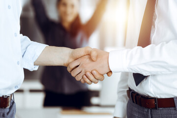 Fototapeta premium Two businessmen shaking hands in sunny office, close-up. Happy and excited business woman stands with raising hands at the background. Business people concept
