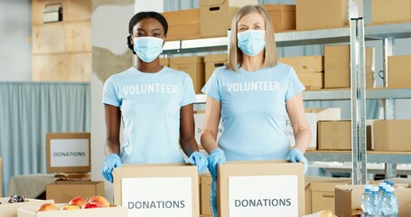 Portrait of positive Caucasian and African American female volunteers in medical masks working in charitable stock organization packing donations sorting food and looking at camera, volunteering