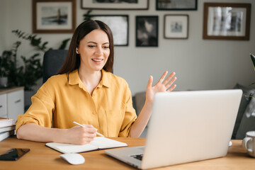 Young pretty woman talking on video call and waving hand while sitting at table in the homeoffice room