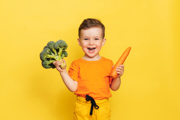 A studio shot of a smiling boy holding fresh broccoli and carrots on a yellow background. The concept of healthy baby food.