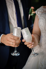 The hands of the newlyweds with gold rings hold white glass glasses with champagne.