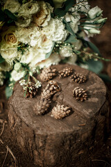 Gold wedding rings on a round wooden stump in the forest, surrounded by fir cones. Nearby, a beautiful bouquet of white and beige roses lies on the ground. Brown, white, beige. Vertical frame.