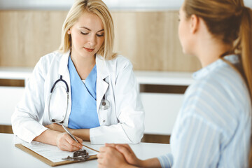 Fototapeta premium Woman-doctor and patient sitting and talking in hospital or clinic. Blonde therapist is cheerfully smiling. Medicine concept