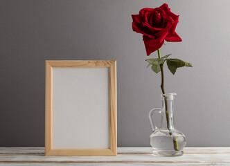 White wooden frame mockup with red rose in glass on gray paper background. still life, copy space.