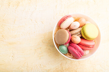 Different colors macaroons and chocolate eggs in ceramic bowl on beige concrete background. top view, copy space.