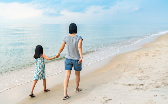 Back view of Asian mother and little daughter are walking forward on the beach, active image with motion blur, perspective view, beautiful sky and sea, blank space for text.