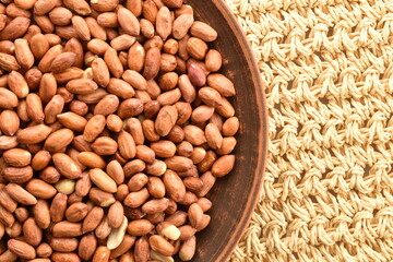 Many grains of organic peeled peanuts in a clay plate, on a straw mat, close-up, top view.
