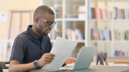Young African Man with Laptop doing Paperwork in Library