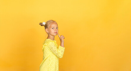 Cute little blonde with bright emotions - attention, mystery, silence, interest. She is wearing a simple yellow dress. Girl's hand is raised, a sign of silence and attention. Yellow background. 