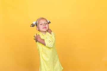 Cute little blonde with bright emotions - surprise, joy, happiness, interest. She is wearing a simple yellow dress.  Girl hugs herself with her hands. Embrace of happiness. Yellow background. European