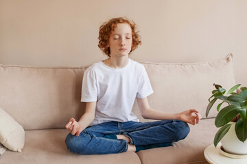 Empty space for text. A teenage boy 14-16 sits on the couch at home and meditates.
