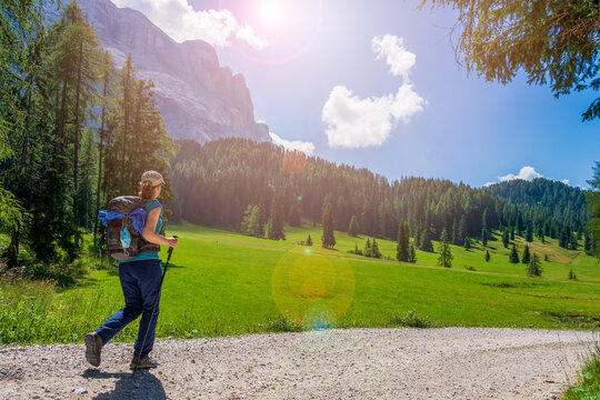Summer: young woman with a hat, trekking poles, a backpack and a face mask attached to it, hiking on a trail through green meadows in the Italian Alps. Dolomite peaks are visible in the background	