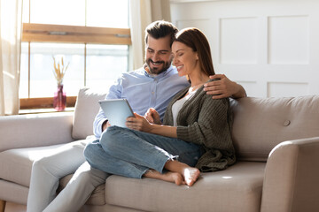 Happy young Caucasian couple relax on couch in living room use modern tablet gadget shopping online. Smiling millennial man and woman renters clients rest on sofa at home talk on video call on pad.