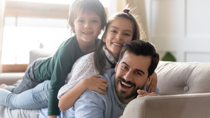 Portrait of smiling young Caucasian single dad and two small kids lying on sofa have fun feeling...