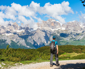 Young man with a hat and a backpack hiking on a trail through green meadows in the Italian Alps. Dolomiti di Brenta mountain peaks are visible in the background. Andalo - Trentino, Italy
