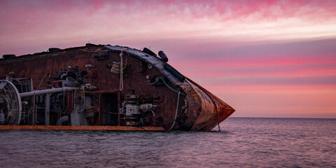 Sunrise time photo of rusty wreaked ship on the purple sea surface below majestic rose and orange clouds.