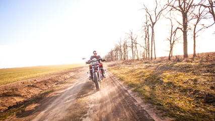 Young curly biker with red glasses drive ride on red chopper through the desert dirt road in late autumn sunset light