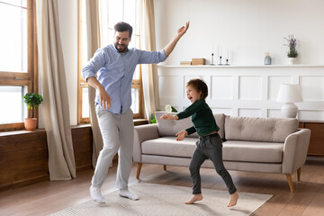 Smiling young father and little 7s son kid dance together feel playful overjoyed on family weekend at home. Happy excited Caucasian dad and small boy child have fun jump play in living room.