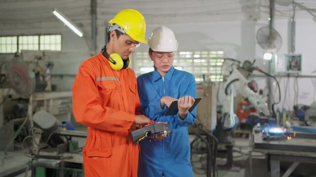 Two engineer wearing uniform and safety helmet standing in the factory and discuss together. Man control robotic arm machine for welding metal and woman holding tablet to check electric system.