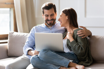 Overjoyed millennial couple relax at home have fun laugh browsing internet on modern computer gadget. Smiling young Caucasian man and woman enjoy weekend use laptop together. Technology concept.