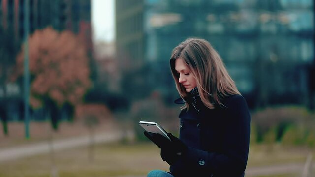 A woman in a black coat sitting in the park works at remote work, using a tablet for work.