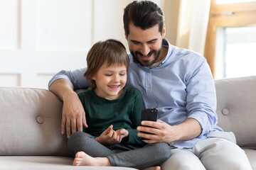 Smiling young Caucasian father and little son sit relax on couch using modern smartphone gadget play together. Happy dad and small boy child browse cellphone talk speak on webcam video call on device.