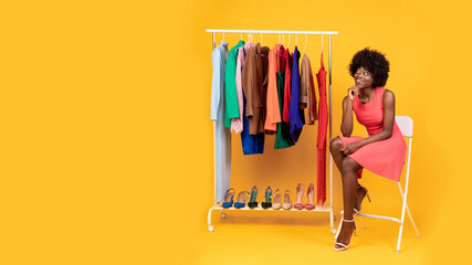 Black Woman Sitting Near Clothing Rack Over Yellow Background