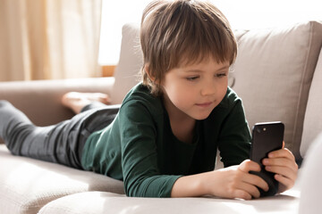 Small 7s Caucasian boy child lying relaxing on sofa in living room use modern smartphone gadget...
