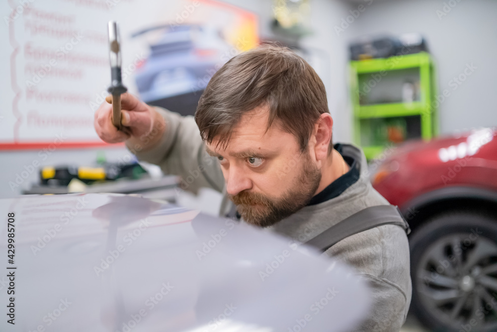 Wall mural Close shot of dent removing without paint, bearded professional worker makes accurate blow rubber hammer 