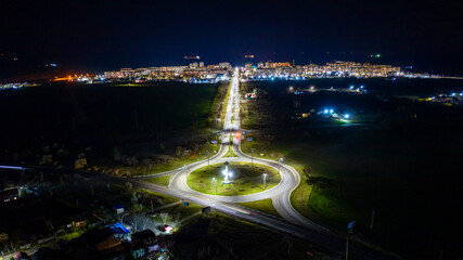 The light on the roundabout at night and movement of cars. Road intersection traffic at night to the bright town on seacoast with ships on background.