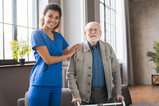 Portrait of young nurse helping senior man to walk using walker equipment in the bedroom. Nurse with old elderly grandfather using walker In rehab center.