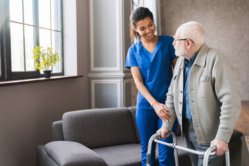 Portrait of an african young nurse helping old elderly disable man grandfather to walk using walker equipment in the bedroom. Senior patient of nursing home moving with walking frame and nurse support - obrazy, fototapety, plakaty