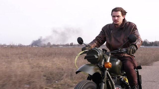 Side view of a stylish cool young man leather jacket driving his vintage motorcycle on an asphalt road with a fire on background in late autumn cloudy evening.