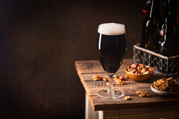 Glass of dark beer with foam head on dark wooden background with empty bottles and beer snacks