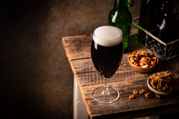 Glass of dark beer with foam head on dark wooden background with empty bottles and beer snacks