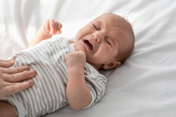 Closeup Portrait Of Crying Little Newborn Baby In Bodysuit Lying On Bed