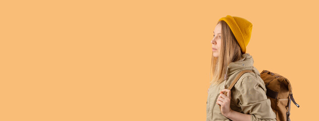 Banner with young girl with long hair in a beige jacket and a yellow hat with a backpack on a solid background, looks away,copy space