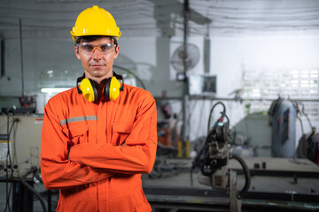 Portrait of young engineer standing and work in the robot factory. Wearing orange uniform, safety helmet and earmuffs crossed arms and smile with copy space. Technology and lifestyle concept