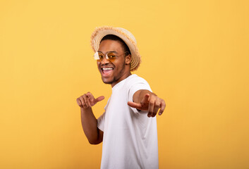 Happy black guy in casual summer outfit, straw hat and sunglasses pointing at you on yellow studio background
