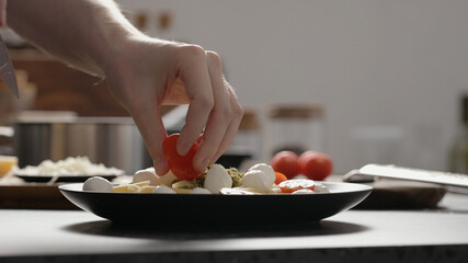 man add cherry tomatoes to pesto penne on black plate