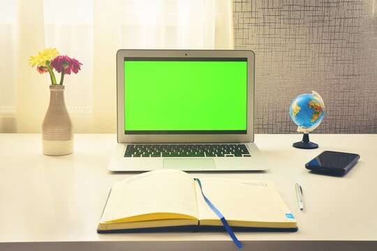 Green screen laptop computer sitting on a home work desk next to notebook, spring flowers and world globe stand. Concept minimalist digital student work space