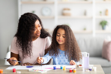 Portrait of happy black mother and her daughter painting with watercolors and gouache at home....