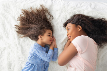 Little black girl with her mom in casual clothes lying on bed together, looking at each other, top view - Powered by Adobe