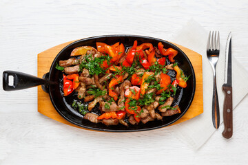 Roast pork with red bell pepper in a frying pan on a white wooden table, knife, fork