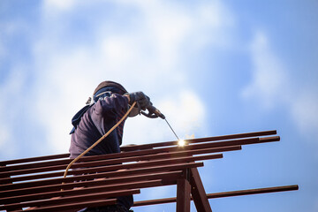 Red painted steel roof truss, with roof welders working at dangerous heights. Welding of steel is very technically demanding and is only professional. Bright sky background