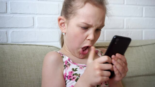 Girl playing mobile game on smartphone on sofa. Preschooler playing mobile phone. Kid using phone for gaming. Child playing video game at home