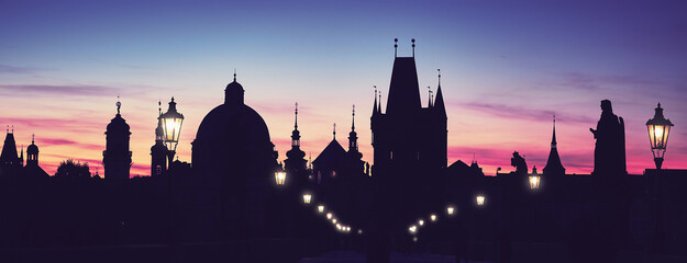 Fototapeta premium Prague, Charles Bridge at night with pink, purple clouds of sunrise. Silhouettes of Old Bridge Tower, churches and spires of Old Prague against light sky with clouds. panoramic banner image.